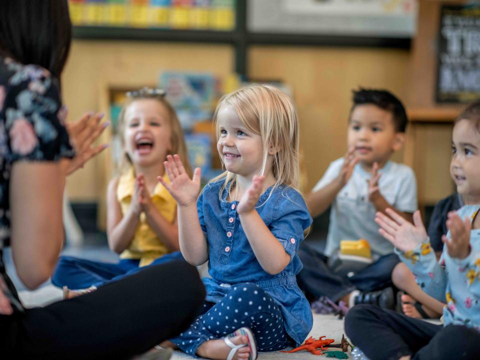 Four preschool-age children seated on the floor playing a singing game with a teacher