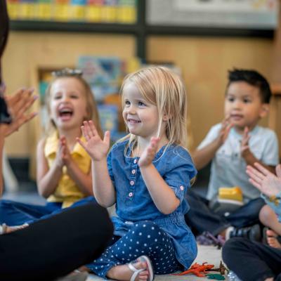 Four preschool-age children seated on the floor playing a singing game with a teacher